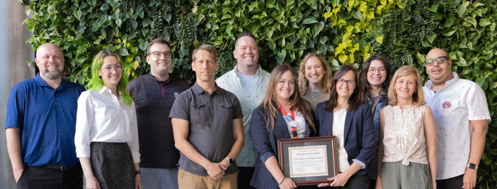 Group of 11 people standing in front of an ivy-covered wall. Two of them are holding a framed certificate.
