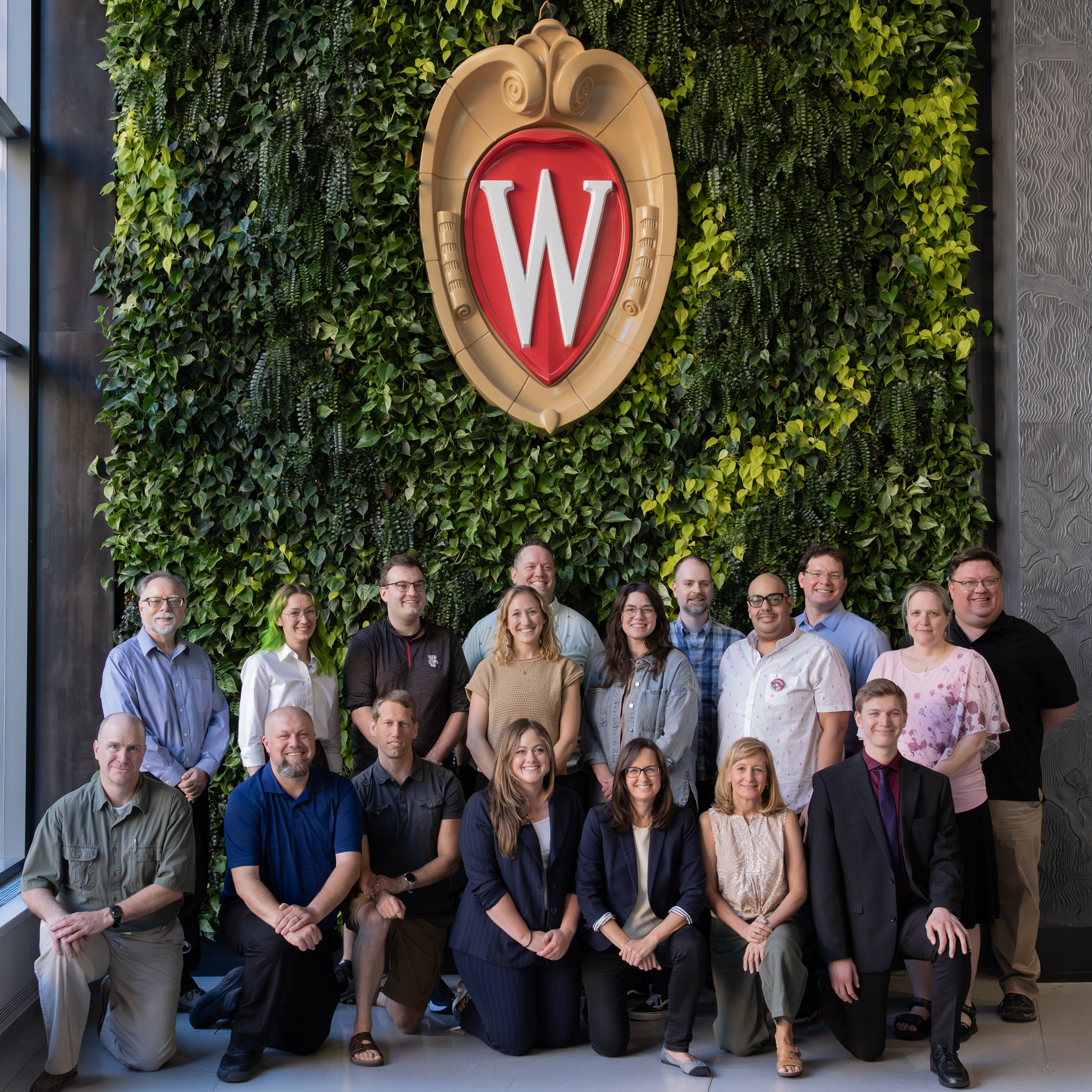 A group of 18 people pose in two rows in front of a living plant wall featuring the UW–Madison "W" crest.