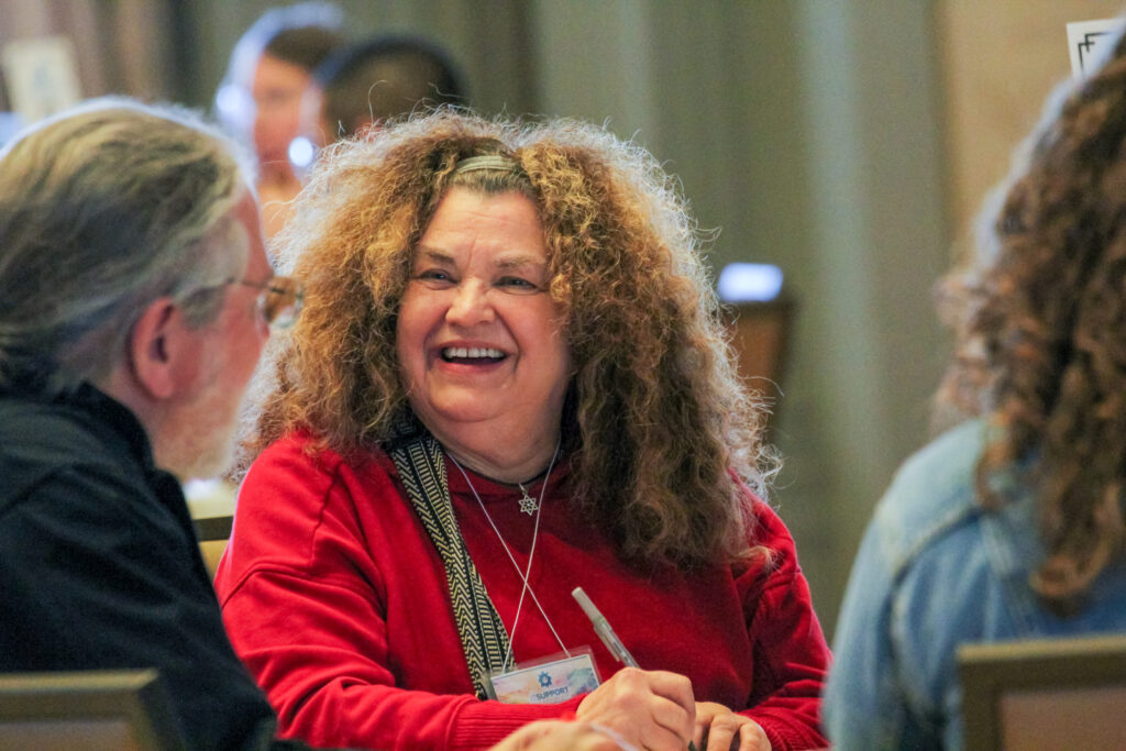 A smiling woman with curly hair and a red sweater laughs during a table discussion.