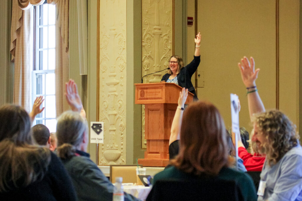 A presenter standing at a podium and audience members raise their hands.