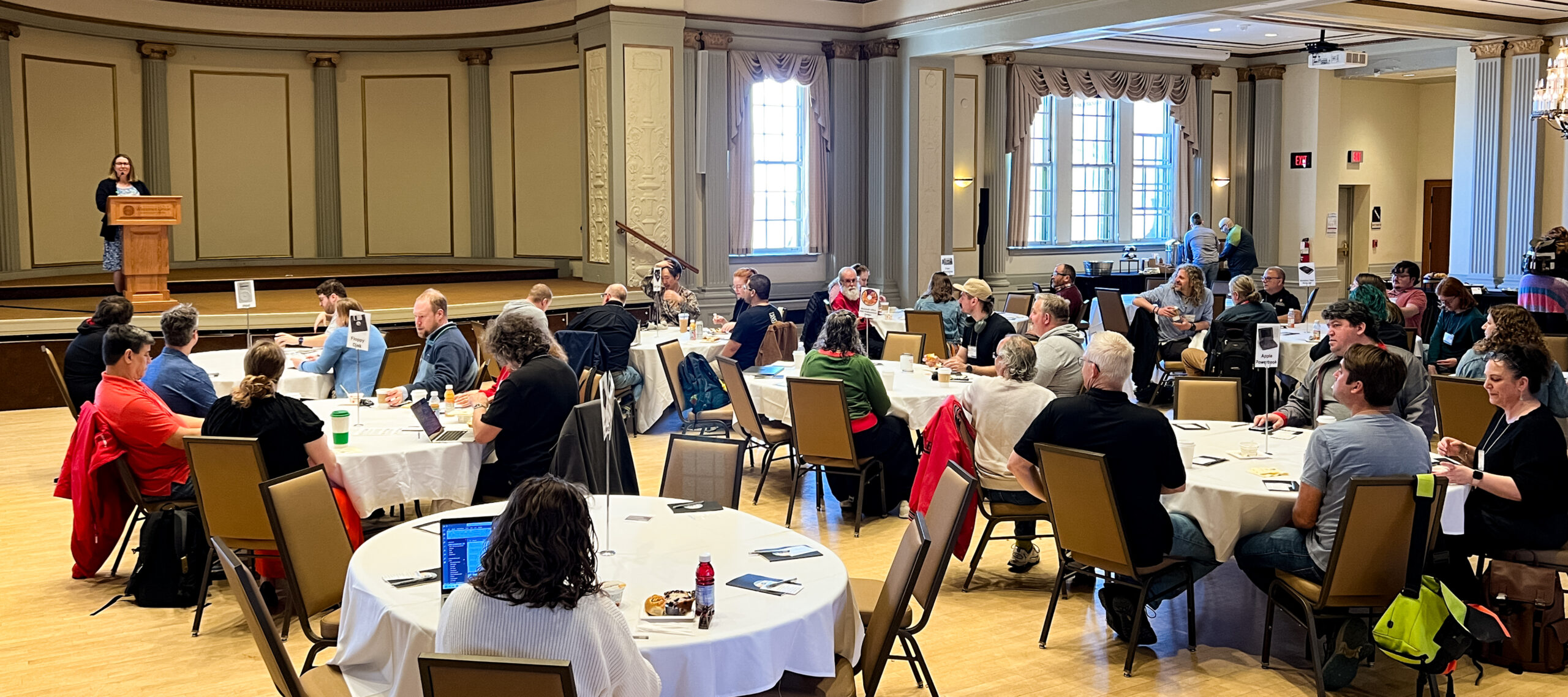 Attendees seated at round tables in a ballroom listen to a presenter at a podium.