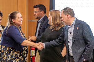 A smiling woman shakes hands with a man in a conference room while others converse in the background.