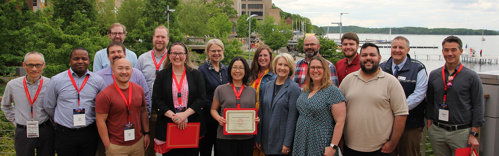 A group of approximately 20 professional colleagues pose together outdoors, with several holding red-framed certificates or awards, and a lake visible in the background.