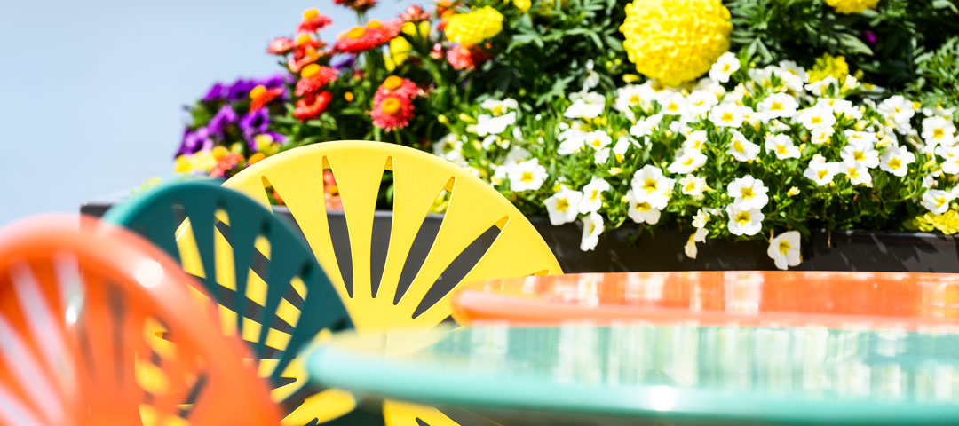 Colorful flowers behind distinctive starburst orange, green and yellow chairs and matching round tables on an outdoor terrace.