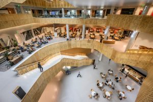 Multi-story atrium of Morgridge Hall showing curved white balconies, perforated golden metal walls, and students gathered at round tables on the ground floor and mezzanine levels.
