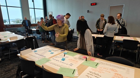 Workshop participants gather around tables with different colored papers and converse.