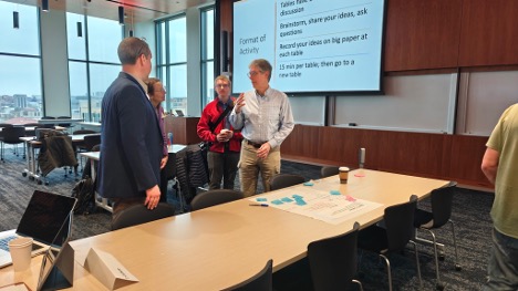 Four people gather in front of a projection screen and next to a table with blue sticky notes.