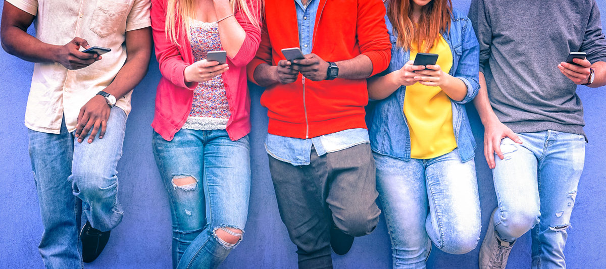 A group of teens standing against a wall using their phones.