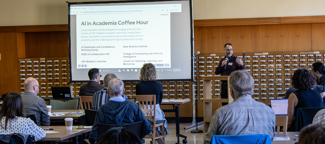 Attendees sit in a conference room while a speaker at a podium presents. The projected slide reads “AI in Academia Coffee Hour” and lists the event’s co-sponsors.