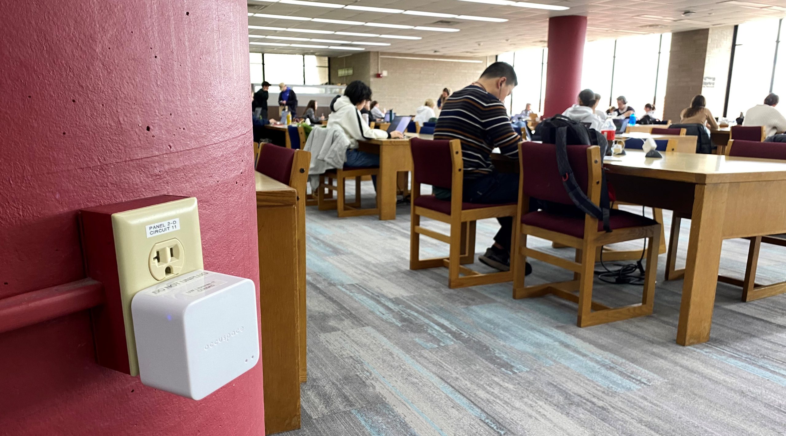 A small, white, square device is plugged into a wall outlet while students study at tables in the background.